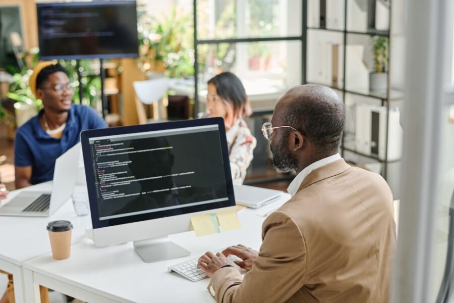 A man types on a computer displaying code related to website maintenance, while two colleagues engage in discussion in the background of a modern office setting.