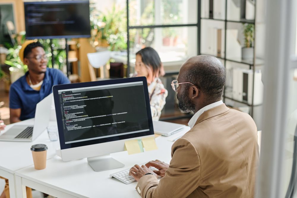 A man types on a computer displaying code related to website maintenance, while two colleagues engage in discussion in the background of a modern office setting.