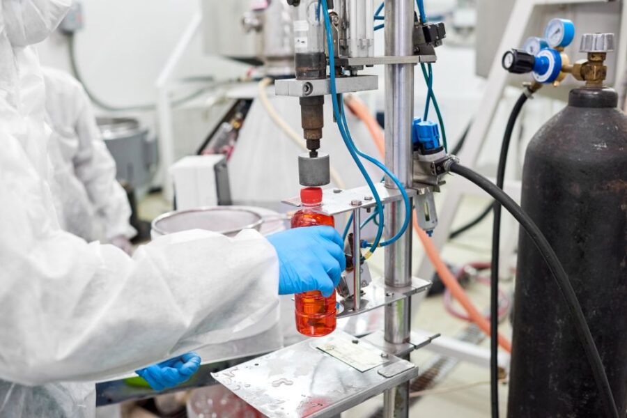 A person in protective clothing operates a filling and capping machine as it fills a plastic bottle with red liquid in a laboratory or industrial setting.