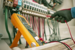 A person performs electrical testing with a digital multimeter, measuring electrical values in a circuit breaker panel filled with multiple colored wires.