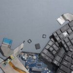 Broken electronic components—including a cracked screen, circuit board, loose keyboard keys, and computer chips—are scattered on a gray surface, highlighting the growing need for proper ewaste recycling.