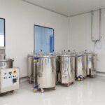 A clean, white room with four large stainless steel industrial tanks on wheels and a control panel, used for processing or storing liquids.