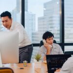 Three people work in a modern office with large windows; one stands by a monitor, another talks on the phone about the Singapore IT firm building grant-eligible security stacks, while a third works on a laptop.