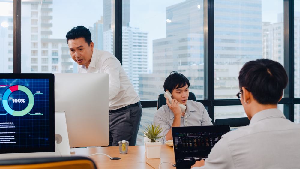 Three people work in a modern office with large windows; one stands by a monitor, another talks on the phone about the Singapore IT firm building grant-eligible security stacks, while a third works on a laptop.